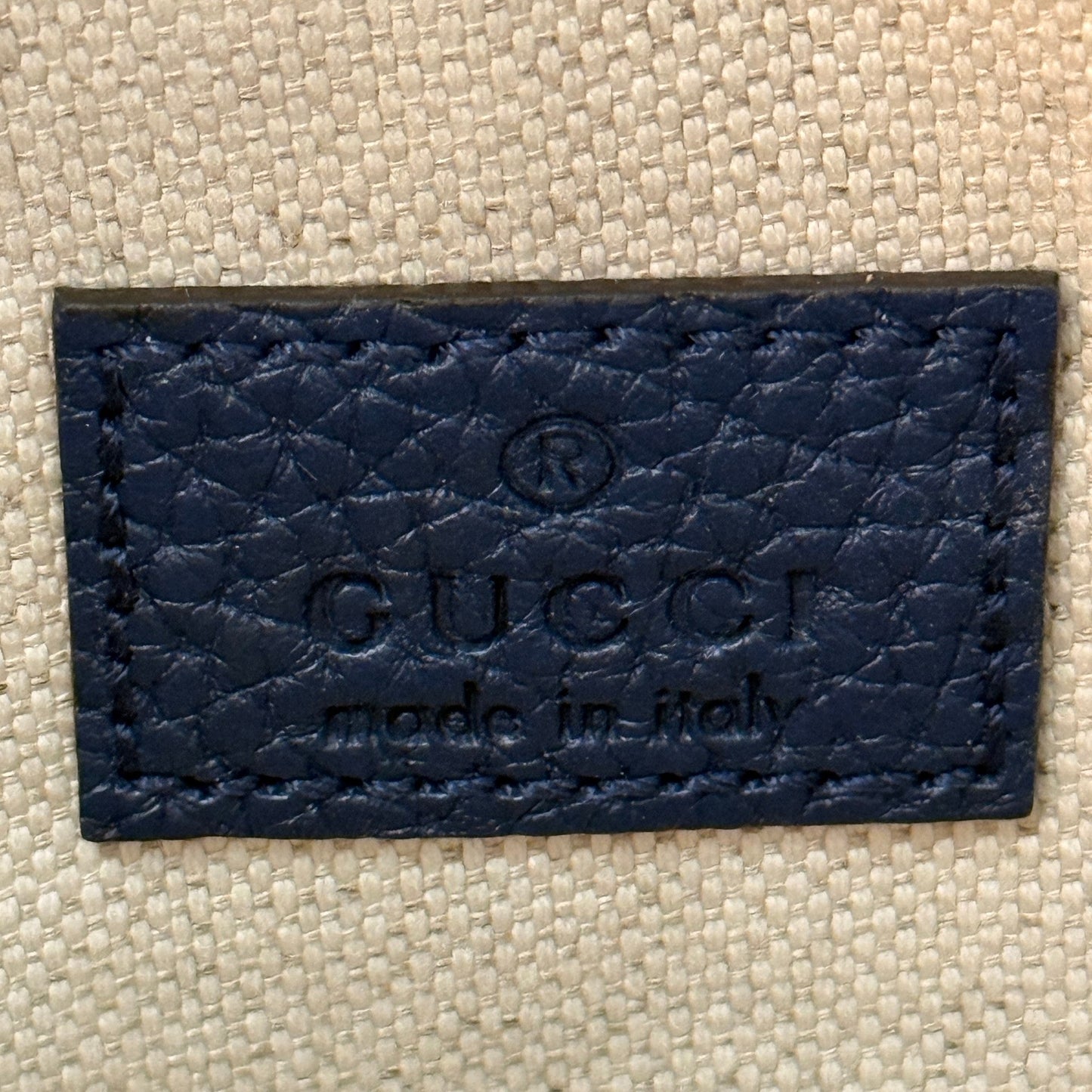 Luxe Vault blue embossed leather Gucci brand tag with Made in Italy stamp on a textured fabric background
